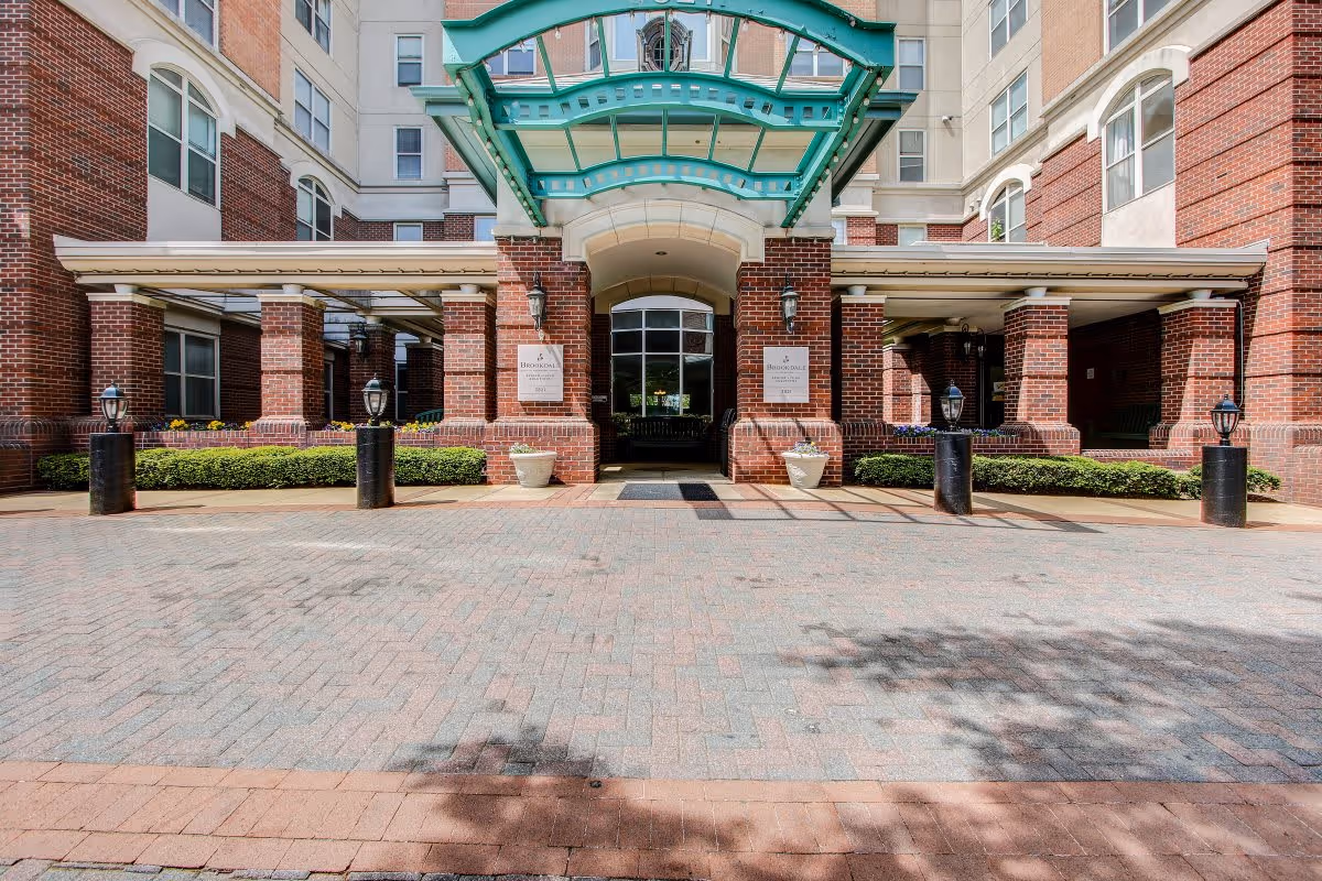 Entrance of a senior living facility with a covered driveway, brick pillars, and a green glass canopy. The building features large windows and well-maintained landscaping with bushes and potted plants near the entrance.