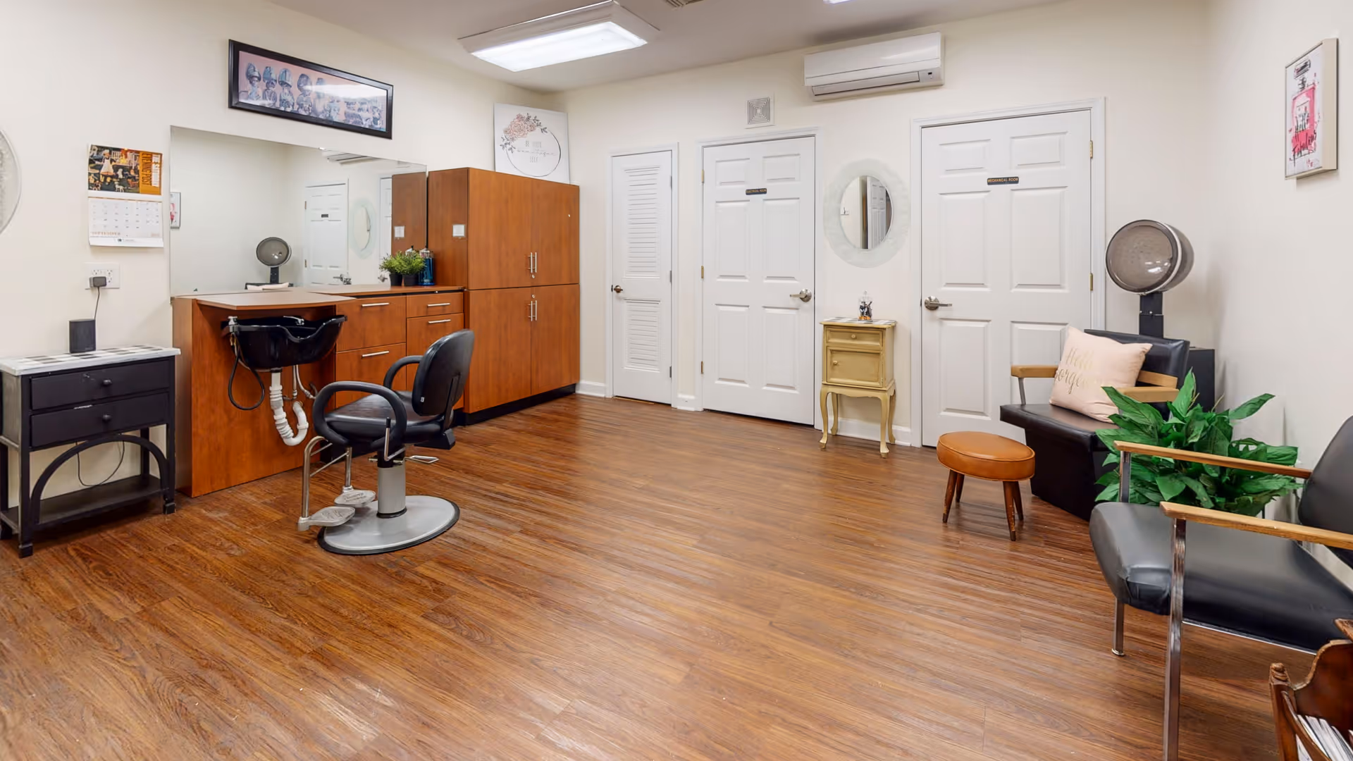 Interior view of a senior living facility's hair salon area with a styling chair in front of a large mirror and wooden cabinetry. The room has wood flooring, white walls, and multiple closed white doors. There are additional chairs, a small table with a plant, and a hair dryer in the corner.