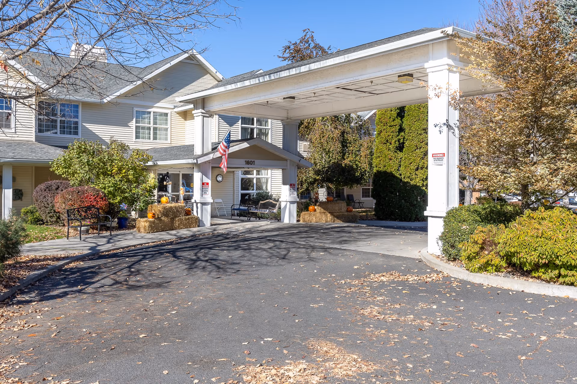 Exterior view of McKay Creek Assisted Living facility entrance with a covered drop-off area, American flag, benches, hay bales decorated with pumpkins, and surrounding trees with autumn foliage.