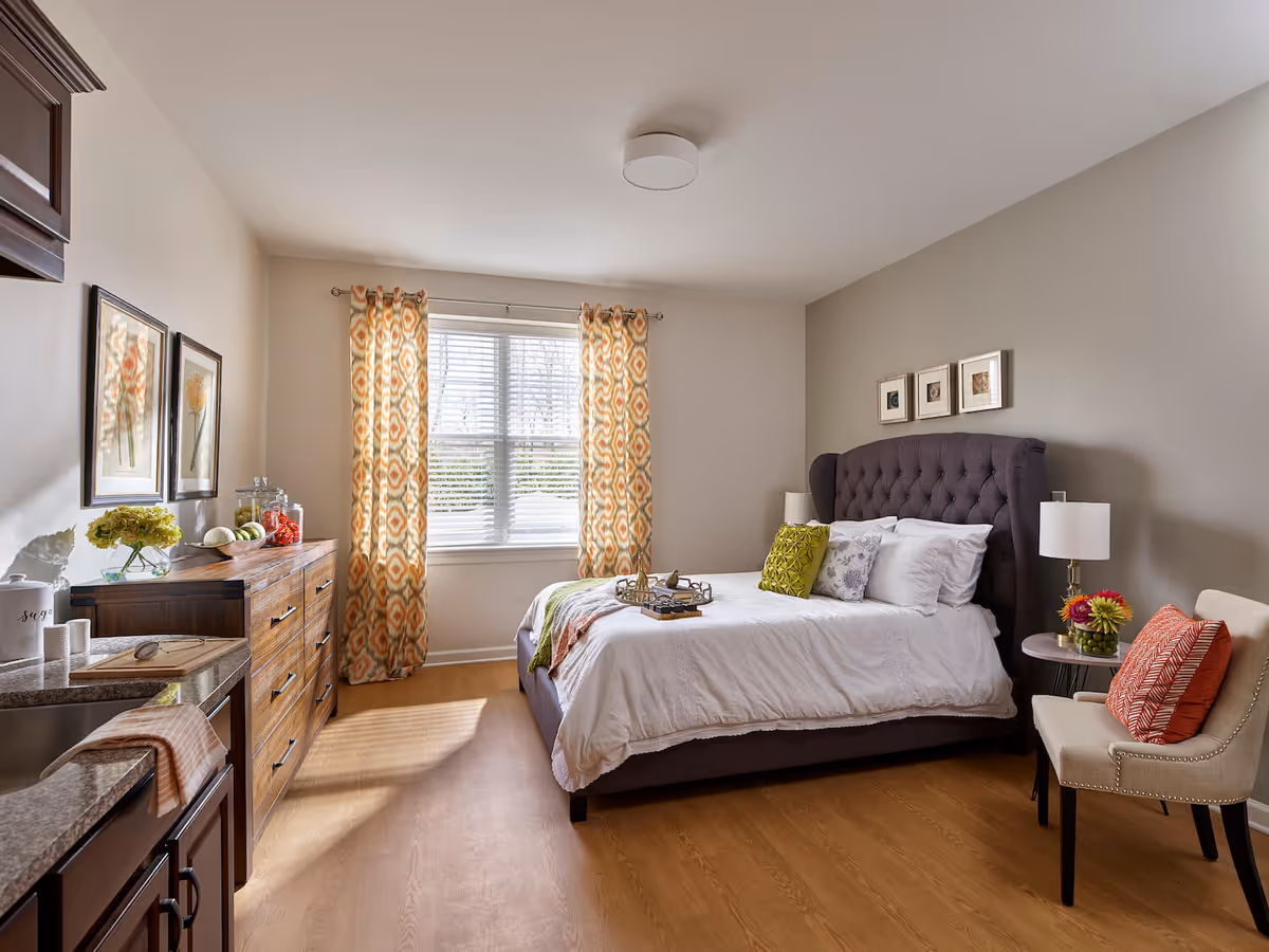 A bright and cozy bedroom in an assisted living facility featuring a large bed with white bedding and decorative pillows, a wooden dresser with glass jars and fruit, a window with patterned curtains letting in natural light, a small side table with a lamp and flowers, and a beige upholstered chair with an orange cushion.