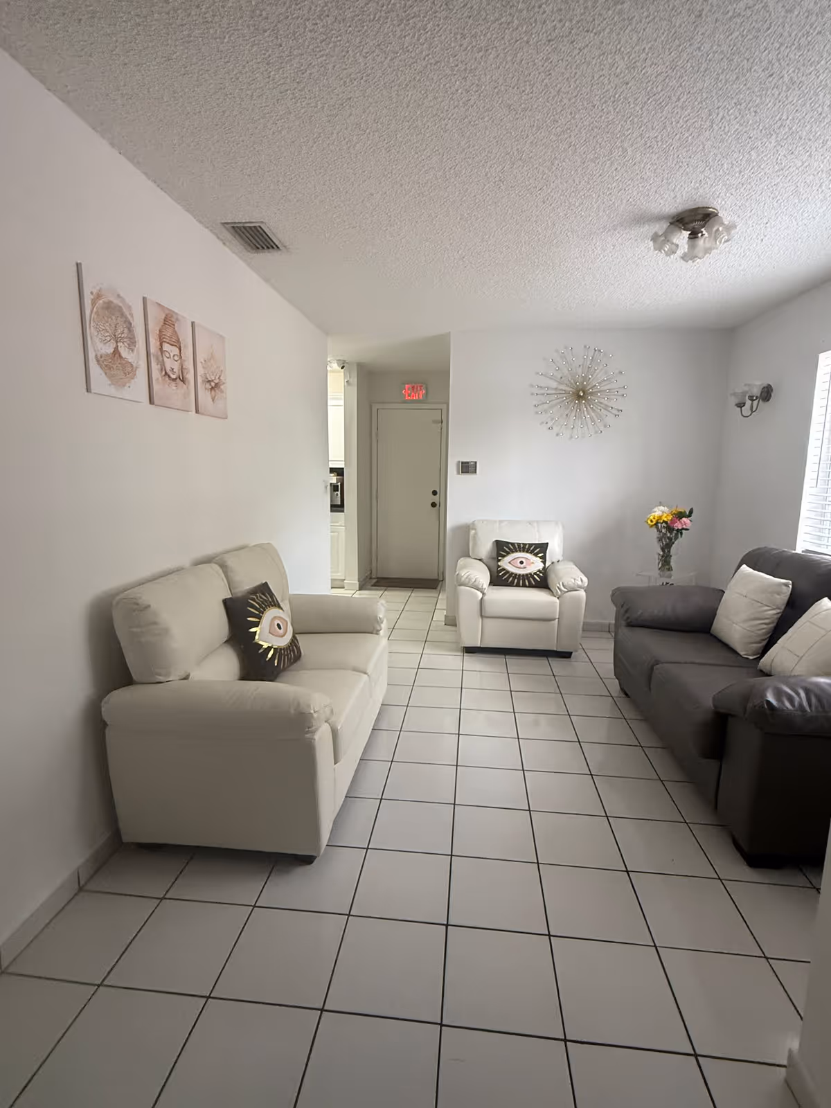 A small living room with white tiled floor and white walls. The room contains a white loveseat with a decorative pillow, a white armchair with a similar pillow, and a dark gray sofa with white pillows. On the wall above the loveseat are three framed artworks, and on the wall above the armchair is a decorative starburst wall clock. A small table with a vase of flowers is next to the armchair. A door with an exit sign above it is visible in the background.