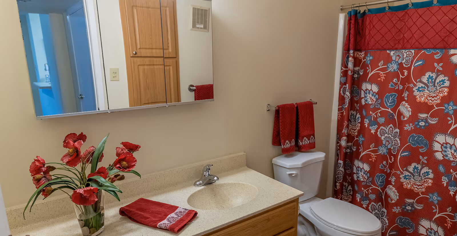 A bathroom with a beige countertop and sink, a wooden cabinet below, a mirror above the sink, a toilet with red towels hanging on a rack above it, and a red floral patterned shower curtain. A vase with red flowers is placed on the countertop.
