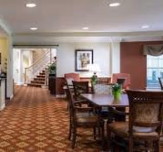 Interior view of a senior living facility common area with a patterned carpet, wooden dining table with chairs, potted plants on the table, armchairs near the wall, a framed picture, a lamp, and a staircase in the background.