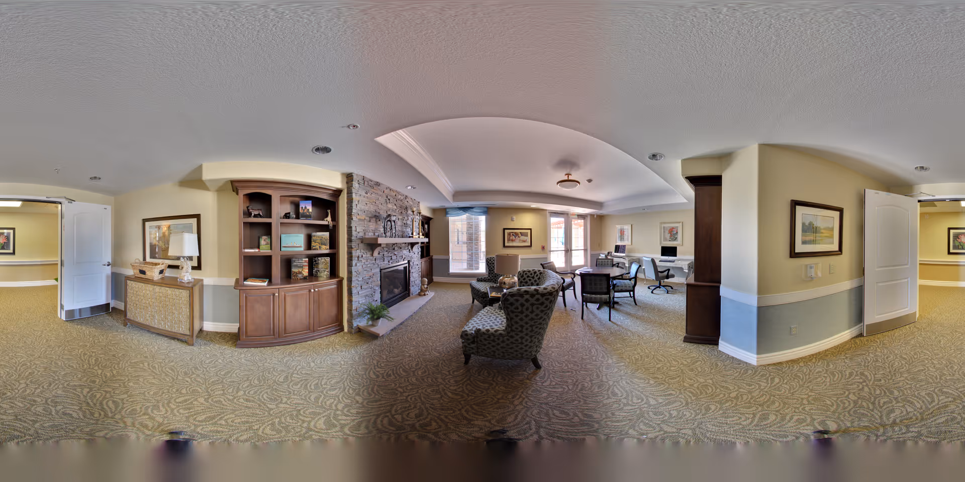 Panoramic common lounge with armchairs, tables, a stone fireplace and built-in shelving in a senior living facility.