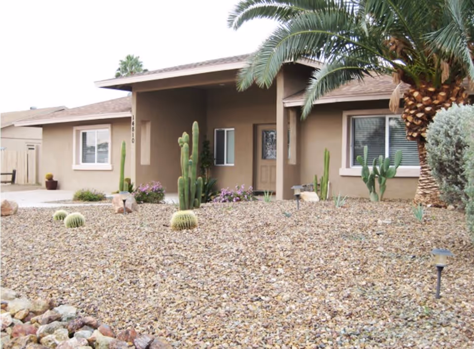 Front exterior view of a single-story building with a covered entrance, desert landscaping with rocks, cacti, and palm trees.