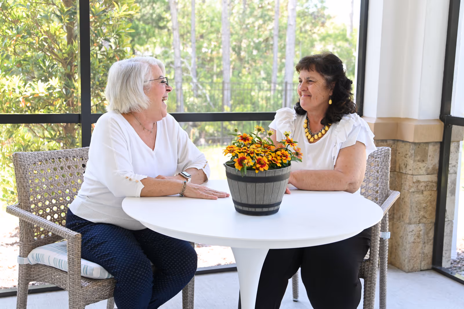 Two elderly women sitting at a round white table on a screened porch, smiling and engaged in conversation. There is a potted plant with yellow and red flowers on the table. The background shows greenery and trees outside the screened area.