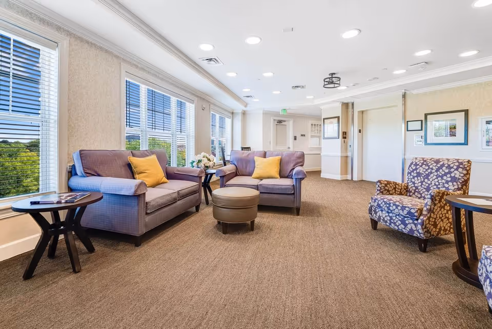 A bright and spacious common area in a senior living facility featuring two purple sofas with yellow pillows, a patterned armchair, a round ottoman, and side tables. Large windows with blinds allow natural light to fill the room, and framed pictures hang on the walls near an elevator.