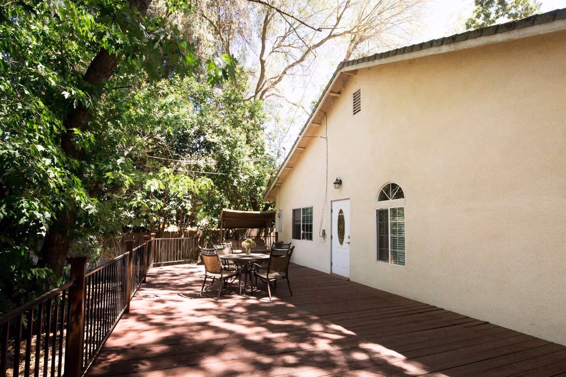 Outdoor wooden deck attached to a beige building with a white door and windows. The deck has a round table with four chairs and a canopy swing. The area is surrounded by green trees and has a black metal railing along the edge of the deck.