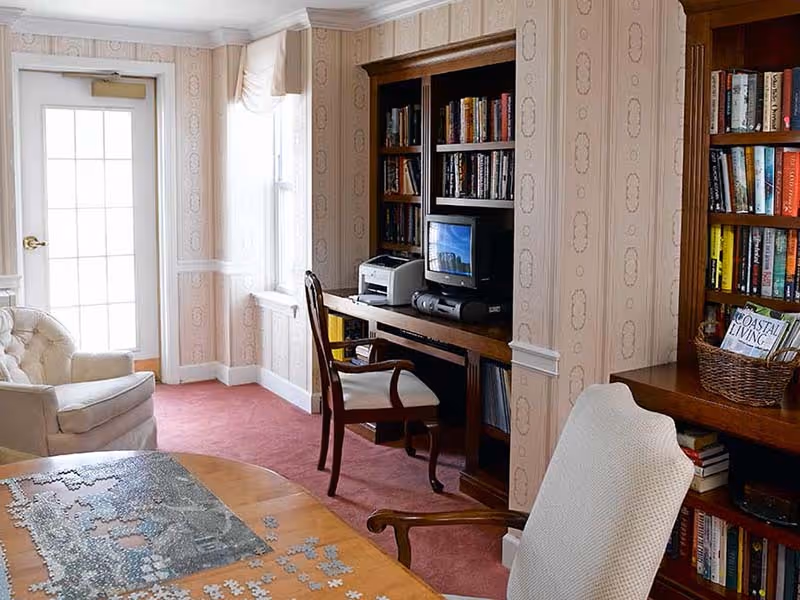 A cozy room with a wooden table partially covered with a puzzle, two upholstered armchairs, a wooden desk with a computer, printer, and bookshelves filled with books. The room has patterned wallpaper, a window with sheer curtains, and a door with glass panes letting in natural light.