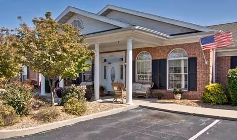 Front view of a single-story brick assisted living facility with porch chairs and an American flag.