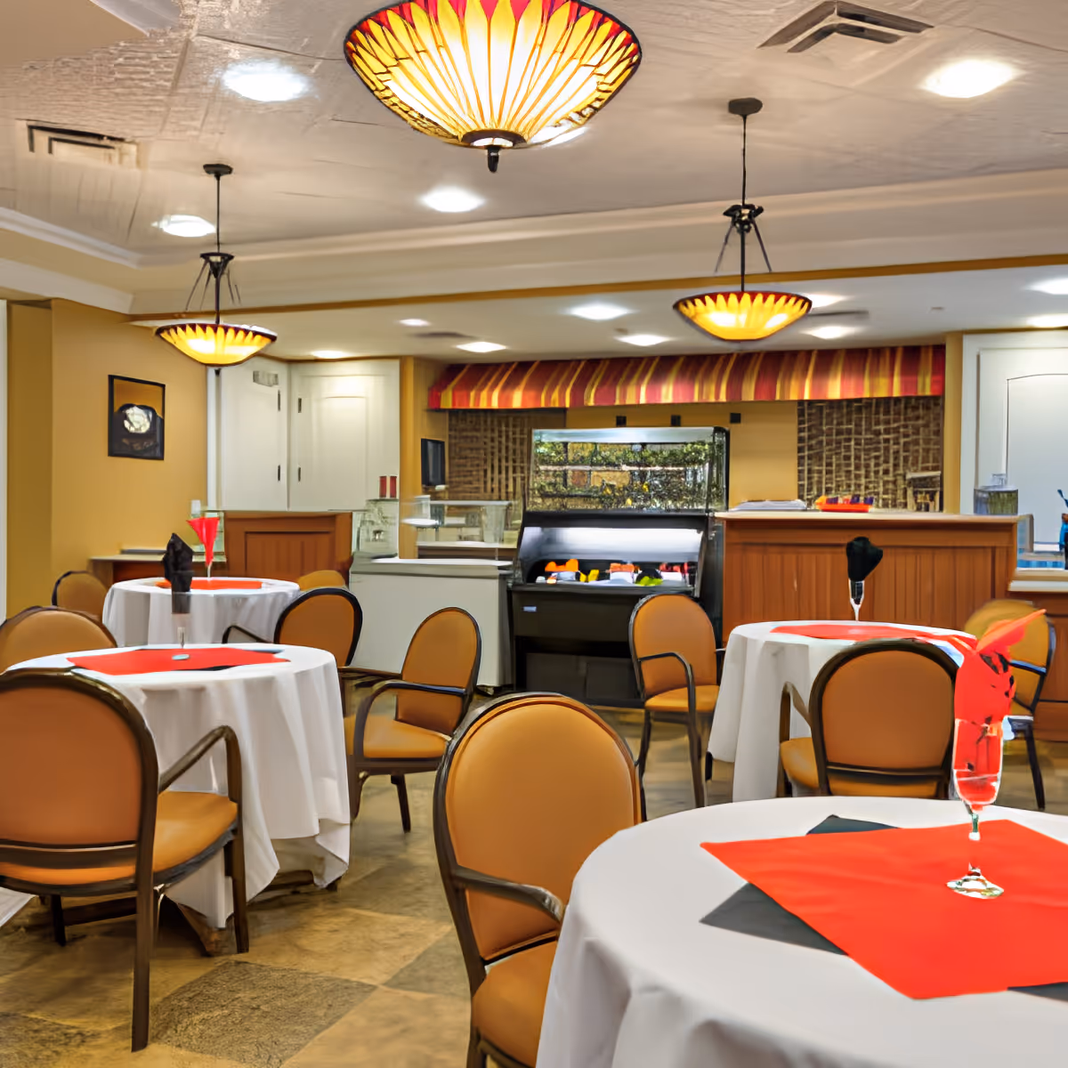 Dining area with round tables covered in white tablecloths and red and black napkins, surrounded by tan cushioned chairs. The room features warm lighting with decorative ceiling lamps and a buffet counter in the background.