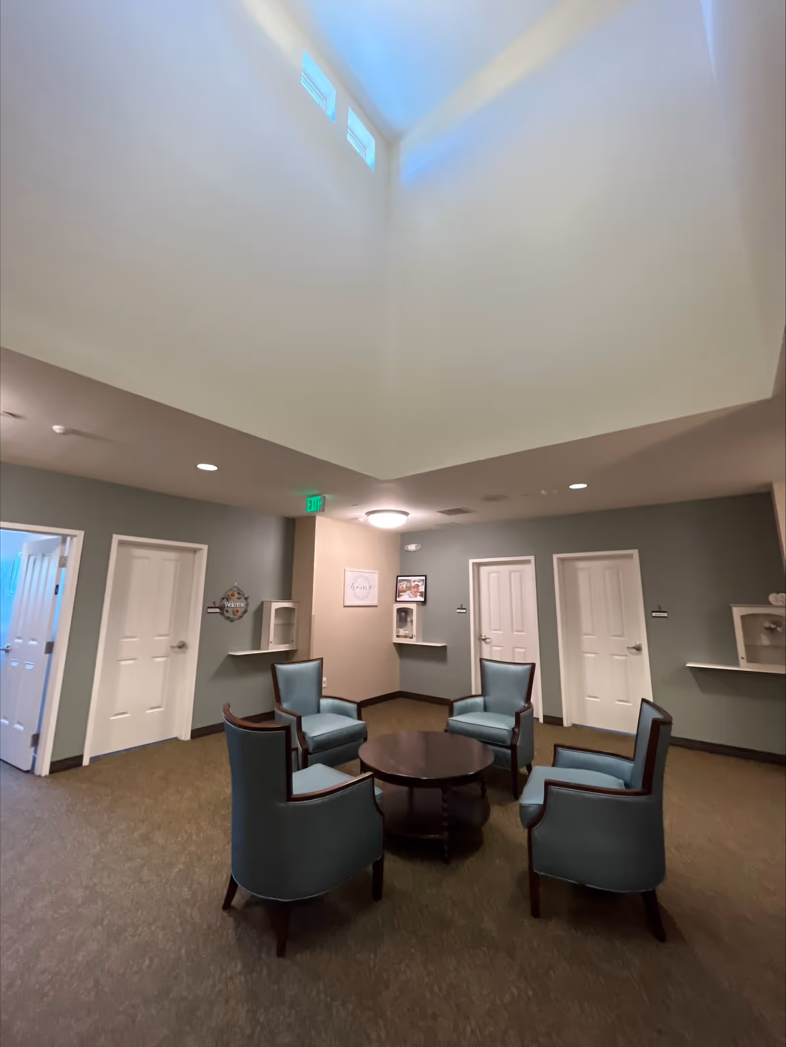 A cozy seating area inside a senior living facility with four blue upholstered armchairs arranged around a round wooden coffee table. The room has beige walls, several white doors, and high ceilings with small windows near the top letting in natural light. There are small wall shelves and framed pictures on the walls.