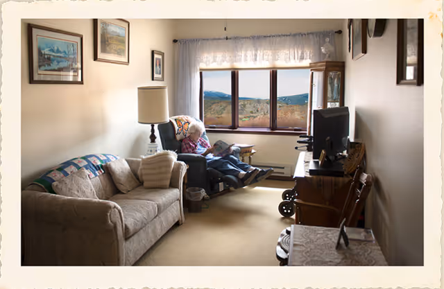 A cozy living room in an assisted living facility with a beige sofa on the left, a standing lamp, and an elderly person sitting in a recliner chair near a large window with a scenic view of hills. The room has framed pictures on the walls, a TV on a wooden stand, and a small table with a lace cover in the foreground.
