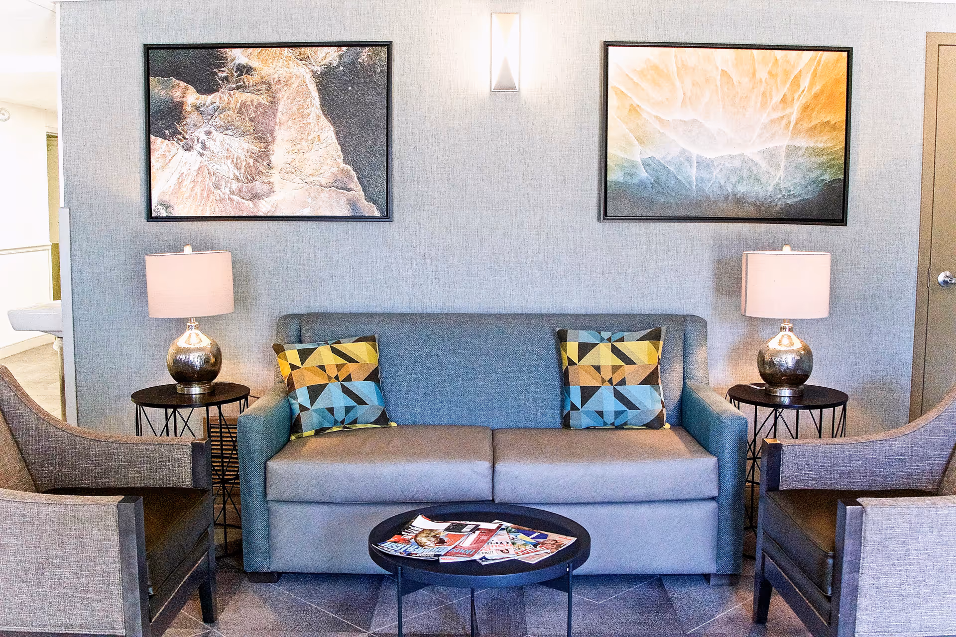 A modern living room area with a blue sofa featuring two geometric patterned pillows, two gray armchairs, two side tables each with a lamp, a round black coffee table with magazines on it, and two abstract paintings on the wall above the sofa.