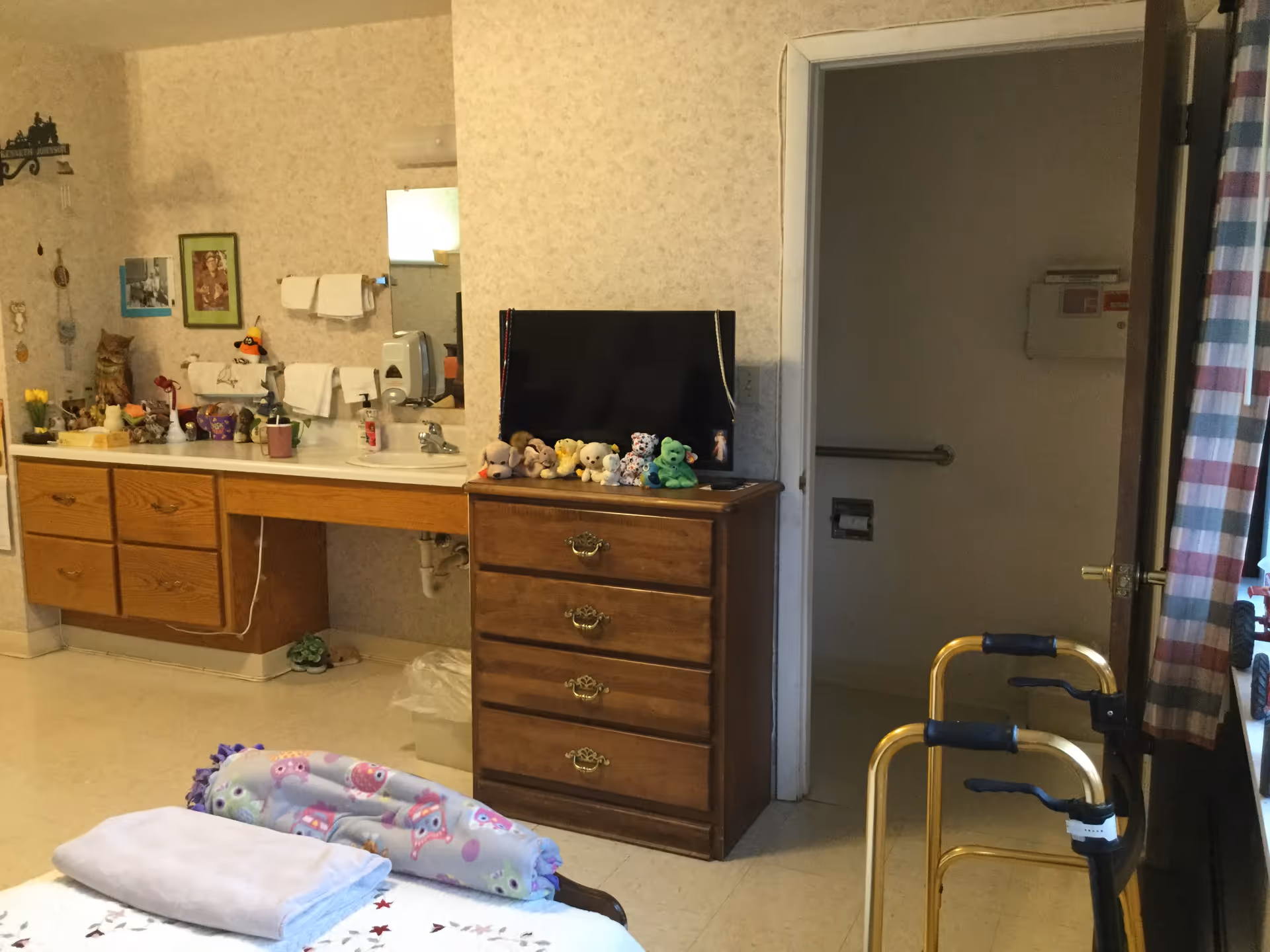 Interior of a senior living facility room showing a bed with folded blankets, a wooden dresser with a TV and stuffed animals on top, a sink with towels and various personal items on the counter, a walker near the window with striped curtains, and an open door leading to a bathroom with grab bars.
