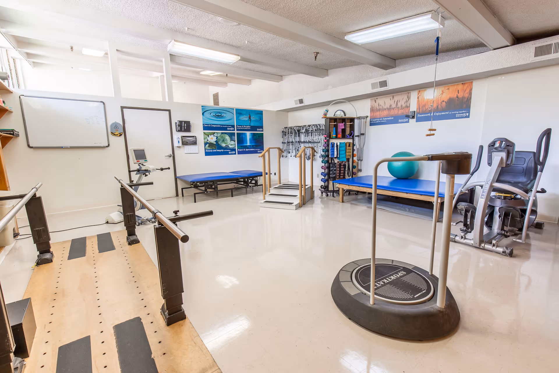 A physical therapy room with various exercise equipment including parallel bars, a balance platform, exercise bikes, therapy tables, and a rack of weights and exercise bands. The room has white walls with motivational posters and a whiteboard.