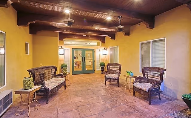 Covered outdoor patio area with wicker seating including two chairs and a loveseat, small side tables, potted plants, and ceiling fans. The patio has a tiled floor and yellow walls with windows and a double glass door leading inside.