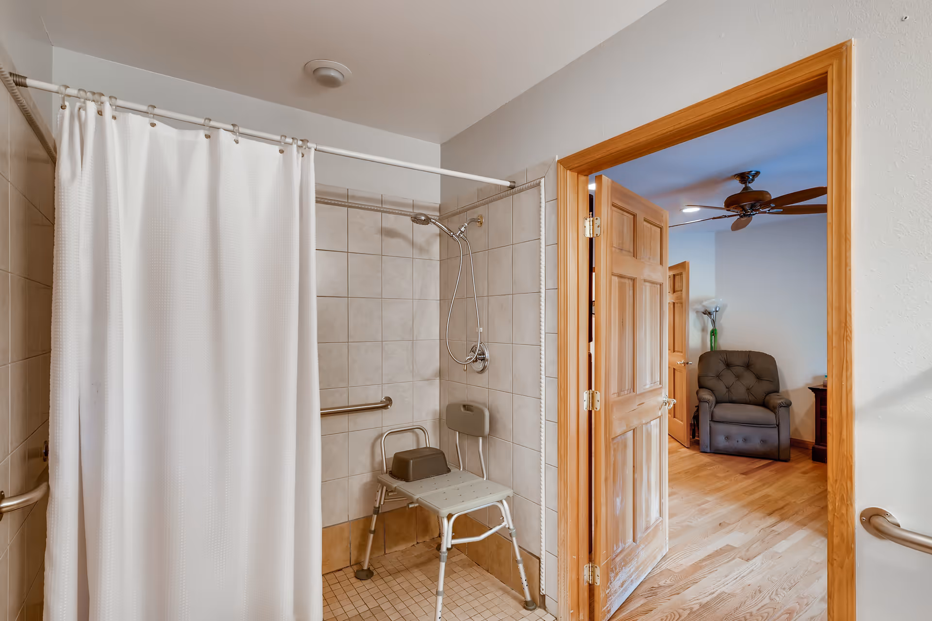 Accessible tiled bathroom with a walk-in shower, shower chair and grab bars, the white curtain partially open to a doorway showing a recliner in the adjacent room.