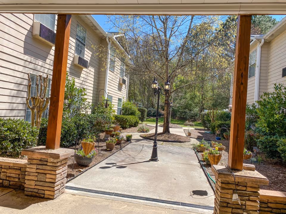 View of an outdoor walkway between two beige buildings with wooden pillars and stone bases. The walkway is lined with potted plants and shrubs, leading to a tree with a vintage-style street lamp. The sky is clear and blue.