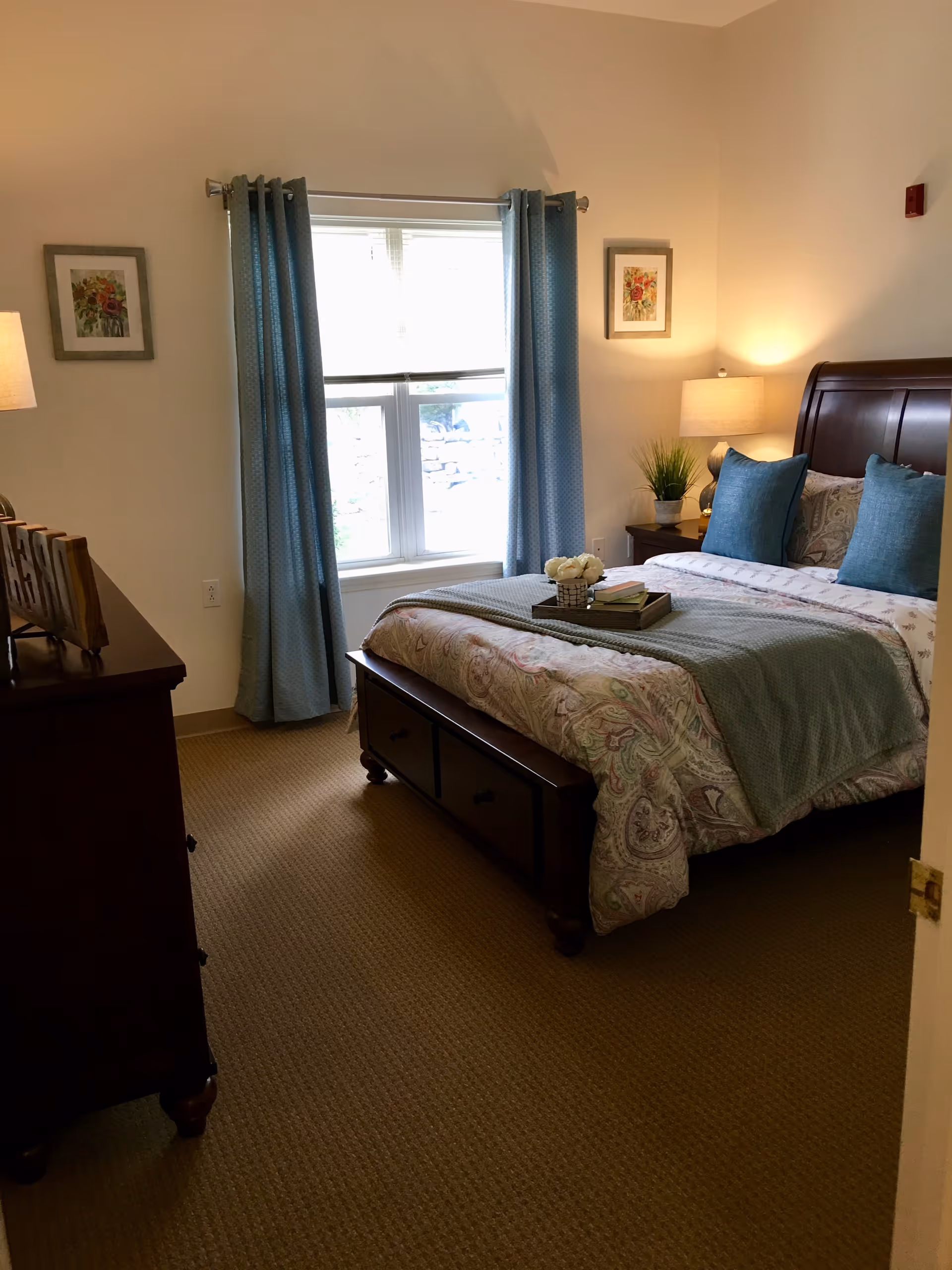 A cozy bedroom with a large bed featuring a paisley-patterned comforter and teal pillows. There is a wooden chest at the foot of the bed, a nightstand with a lamp and a small plant, and a dresser with decorative items. The room has beige carpeting, light-colored walls, two framed floral pictures, and a window with blue curtains letting in natural light.