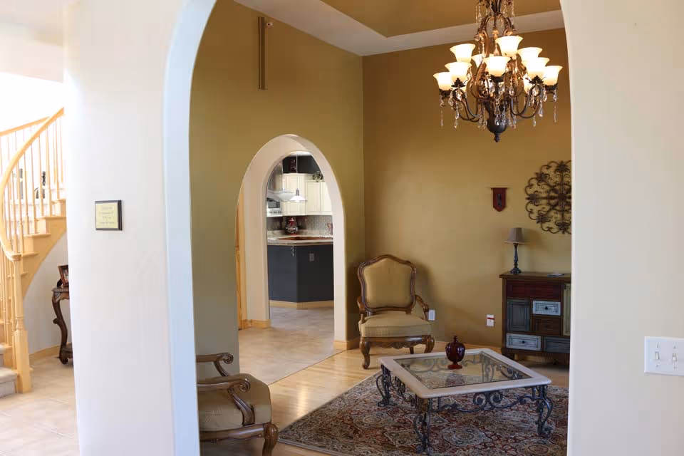 Interior view of a living room area with a decorative chandelier, two upholstered armchairs, a glass-top coffee table on a patterned rug, and a small wooden cabinet against a mustard-colored wall. An arched doorway leads to a kitchen area with white cabinets and a tiled floor. A staircase with wooden railings is visible to the left.