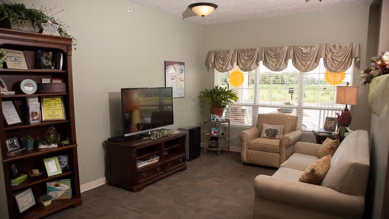 A cozy living room in Providence Place Memory Care featuring a beige sofa with two pillows, a matching armchair with a decorative pillow, a wooden TV stand with a flat-screen television, a tall wooden bookshelf filled with framed photos and decorative items, a small side table with a plant, and large windows with beige valances letting in natural light.