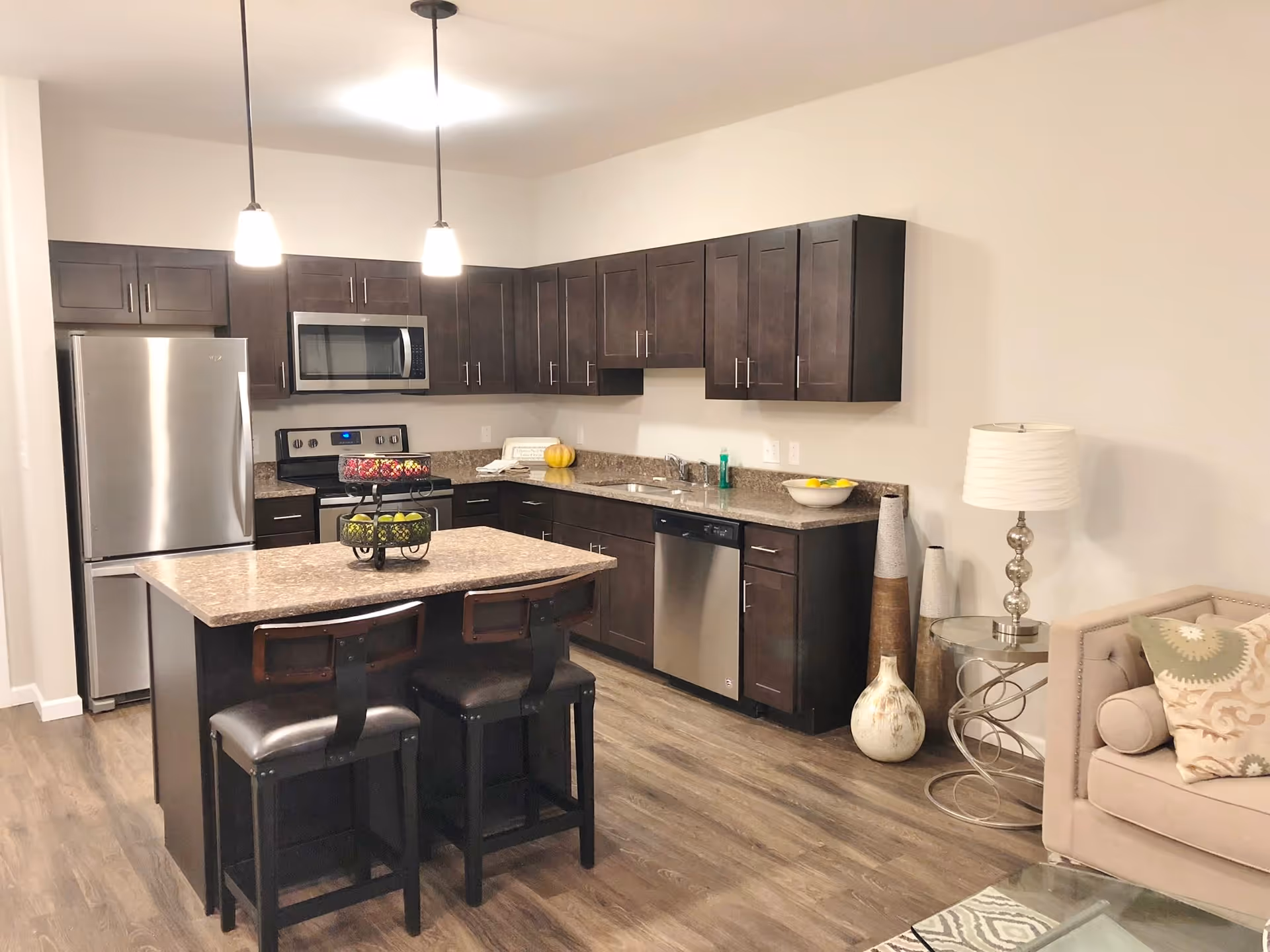 Modern kitchen with dark wood cabinets, stainless steel refrigerator, microwave, stove, and dishwasher. A kitchen island with two chairs is in the center, topped with a two-tiered fruit basket. To the right, part of a beige sofa with patterned pillows and a glass side table with a lamp and decorative vases are visible.