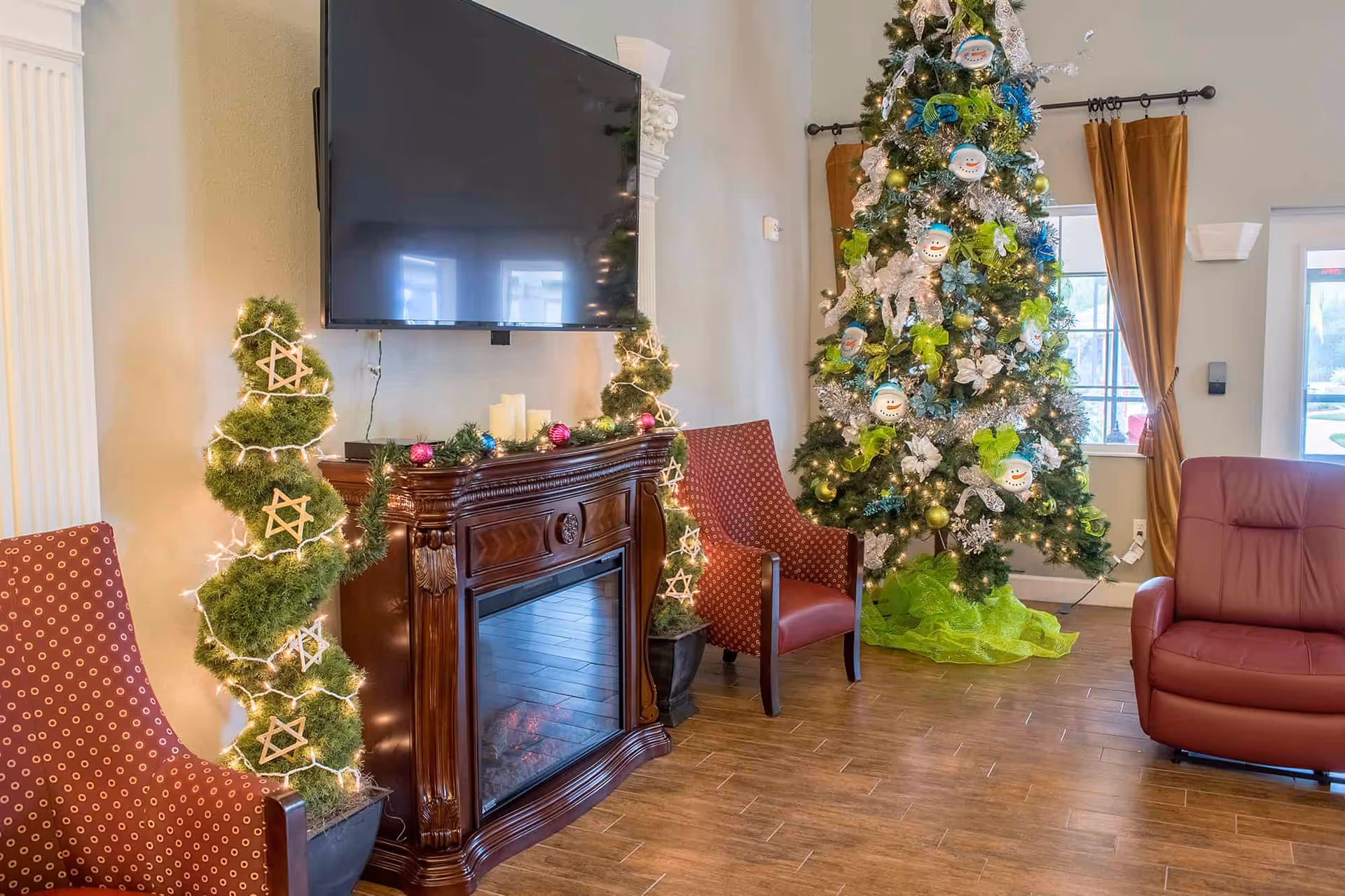 A cozy living room area decorated for Christmas with a large decorated Christmas tree, two red patterned armchairs, a red recliner, a wooden electric fireplace with candles on top, and a flat-screen TV mounted on the wall above the fireplace. There are also two spiral-shaped green plants wrapped with string lights and star ornaments on either side of the fireplace.
