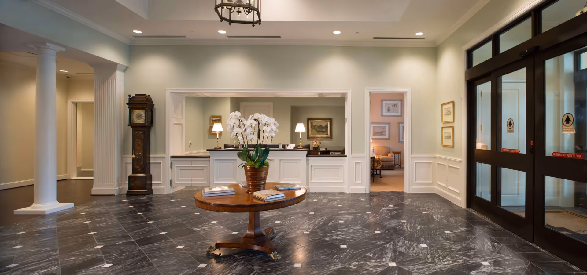 Lobby area of The Tradition–Woodway featuring a round wooden table with a potted white orchid and several books on it, a reception desk with two lamps behind it, a grandfather clock to the left, and glass automatic doors to the right. The floor is covered with dark marble tiles with white accents, and the walls are painted light green with white wainscoting.