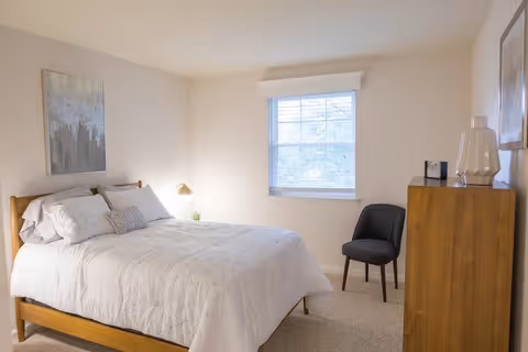 A bright bedroom with a wooden bed frame and white bedding, a window with blinds letting in natural light, a wooden dresser with decorative items on top, a black chair, and a framed artwork on the wall.