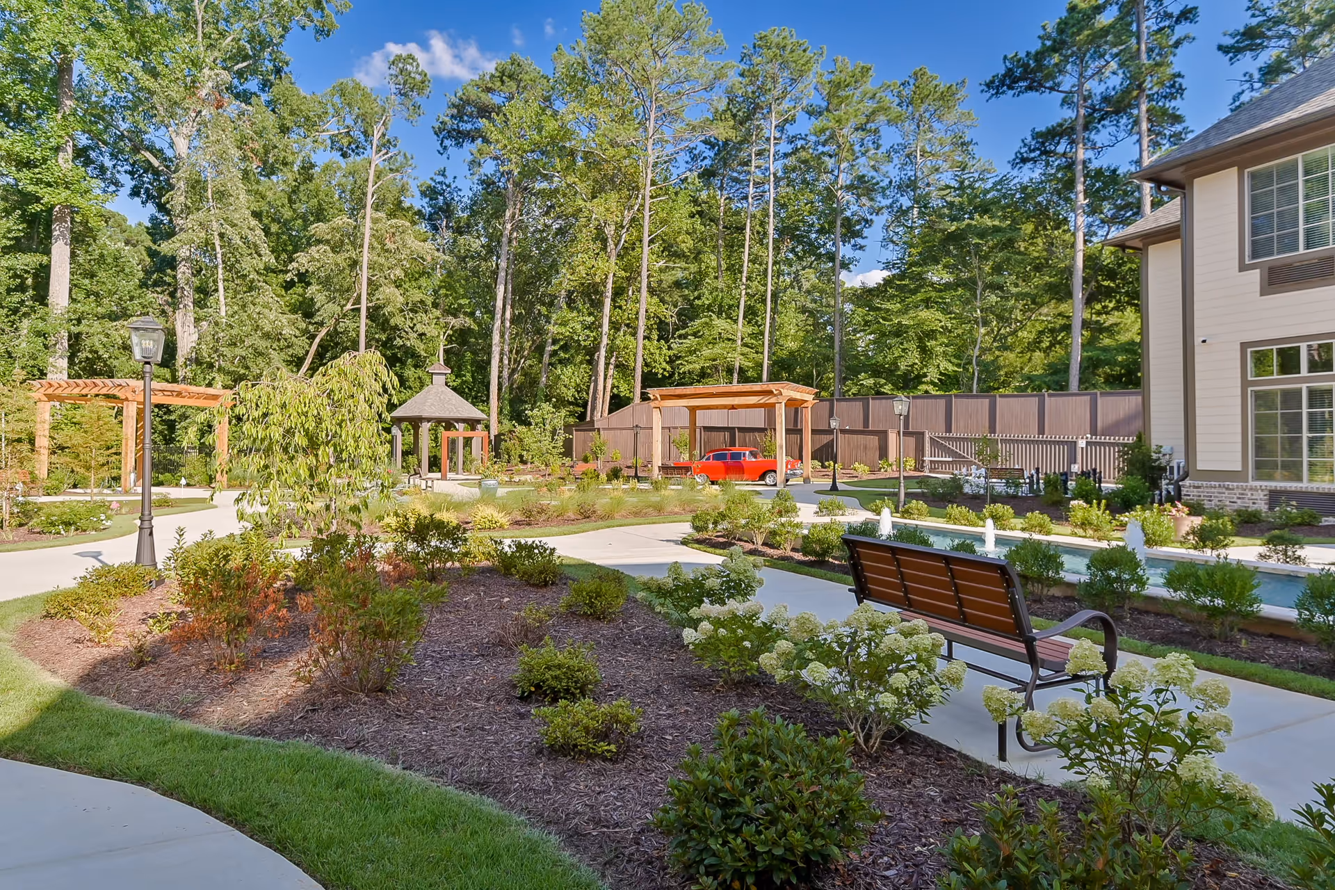 A landscaped outdoor garden area at Orchard at Brookhaven featuring a paved walkway, a wooden bench, a small water fountain, various shrubs and flowering plants, tall trees in the background, a wooden pergola, a gazebo, and a red car parked near a wooden fence.