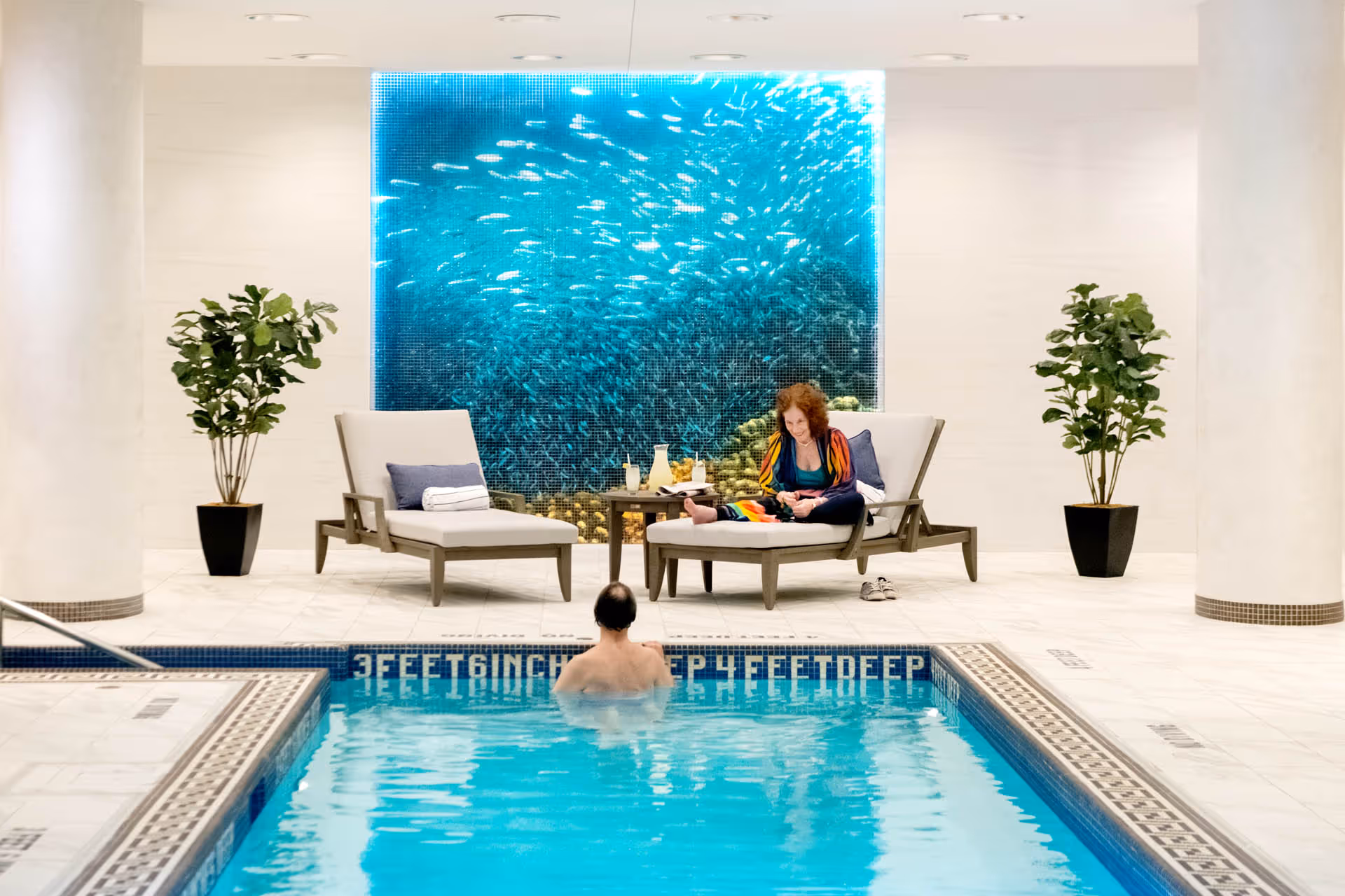 Indoor swimming pool area with a person in the pool facing away and a woman sitting on a lounge chair beside the pool. The woman is wearing a colorful robe and is looking at something in her hands. There are two potted plants on either side of the lounge chairs and a large decorative panel on the wall behind them depicting a school of fish underwater.