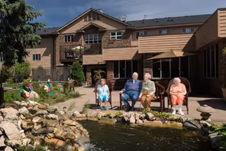Four elderly people sitting on chairs by a pond with rocks and a small waterfall in an outdoor courtyard area of a senior living facility with a multi-story brick building in the background under a clear sky.