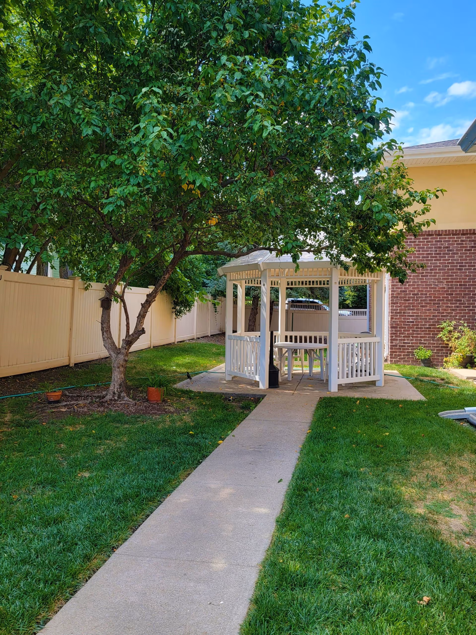 A peaceful outdoor garden area with a concrete pathway leading to a white gazebo. The gazebo has a table and chairs inside. There is a large leafy tree on the left side and a brick building on the right. The area is surrounded by a white fence and green grass.