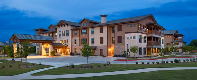 Exterior view of a large, modern senior living facility building named Isle at Cedar Ridge during twilight, with well-lit entrances, multiple windows, balconies, and surrounding landscaped greenery and paved walkways.