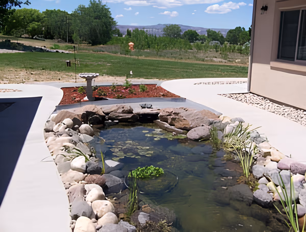 Outdoor view of a landscaped garden area at Grandview Care Lodge Assisted Living featuring a small pond surrounded by rocks and plants, with a concrete pathway and green lawn in the background under a blue sky with some clouds.