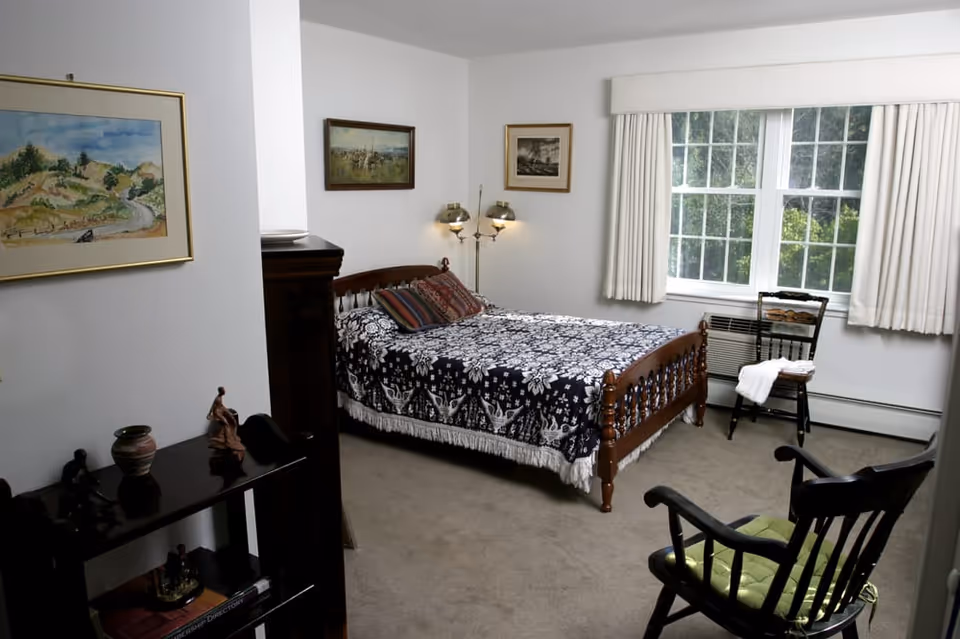 A cozy bedroom with a wooden bed covered in a black and white patterned blanket and several pillows. The room has beige carpet, a window with white curtains, a wooden chair with a white towel draped over it, and a rocking chair with a green cushion. There are three framed paintings on the white walls and a small dark wooden shelf with decorative items.