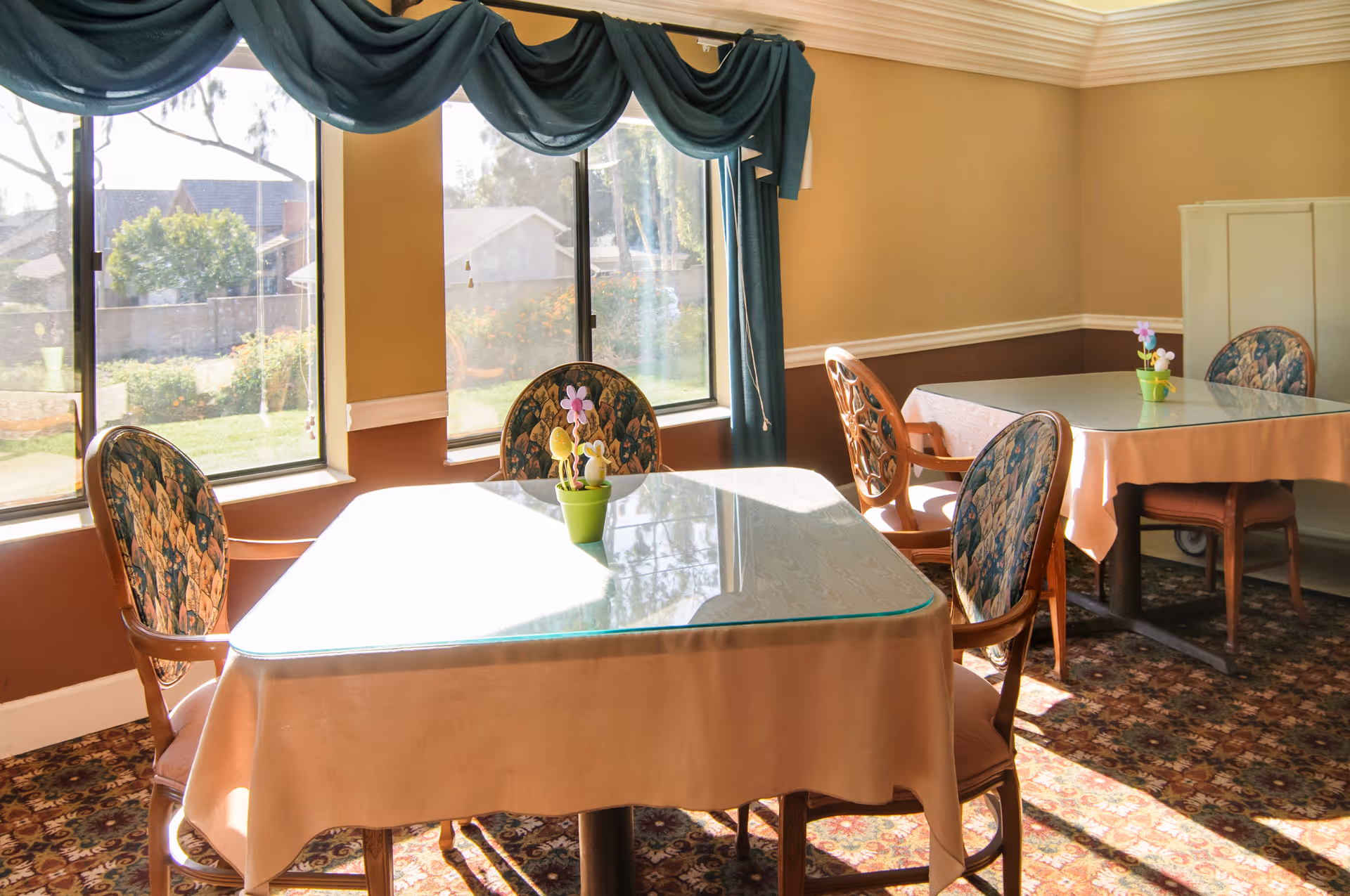 A bright dining room with two tables covered with beige tablecloths and glass tops. Each table has four wooden chairs with floral patterned upholstery. Small flower pots with decorative flowers are placed on the tables. Large windows with teal curtains allow natural light to fill the room, and a patterned carpet covers the floor.