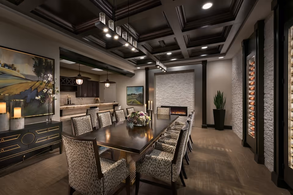 A spacious, elegant dining room with a long dark wooden table surrounded by twelve upholstered chairs with patterned fabric. The room features a coffered ceiling with recessed lighting and two modern pendant lights above the table. On one side, there is a fireplace set into a white textured stone wall, a tall plant in a black pot, and a glass-enclosed wine rack. On the opposite wall, there is a sideboard with candles and a large landscape painting. The room has a warm, inviting atmosphere with neutral tones and sophisticated decor.
