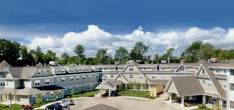 Large multi-wing senior living facility building with a covered entrance, driveway, parked cars, and landscaped grounds under a blue sky.