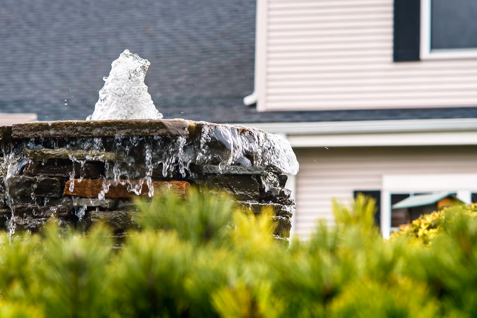 Close-up view of a stone water fountain with water flowing over the edges, surrounded by green shrubs, with a residential building in the background.