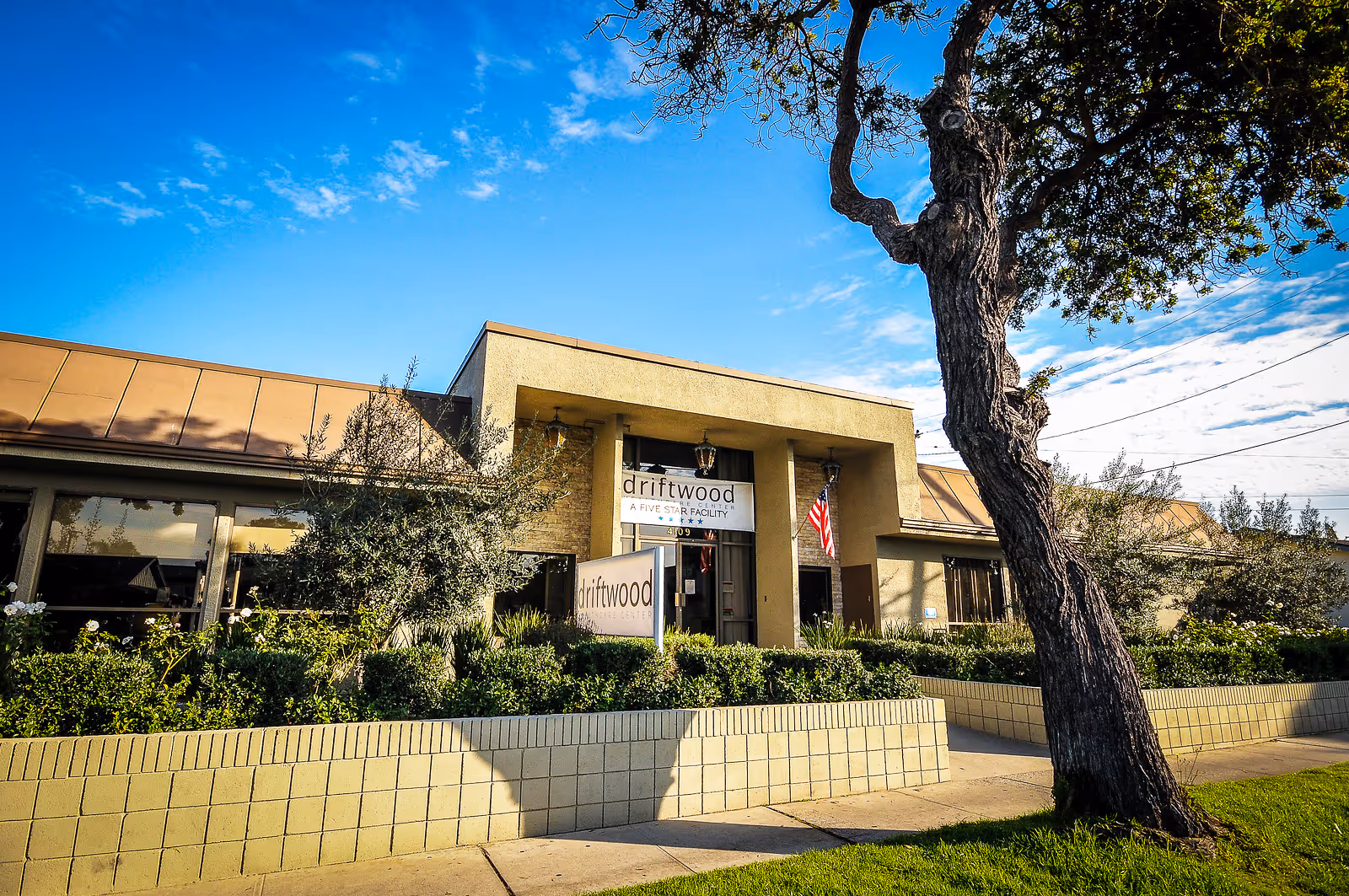 Exterior view of Driftwood Healthcare & Rehabilitation Center building with a beige facade, large windows, an American flag near the entrance, and a tree in the foreground under a blue sky with some clouds.