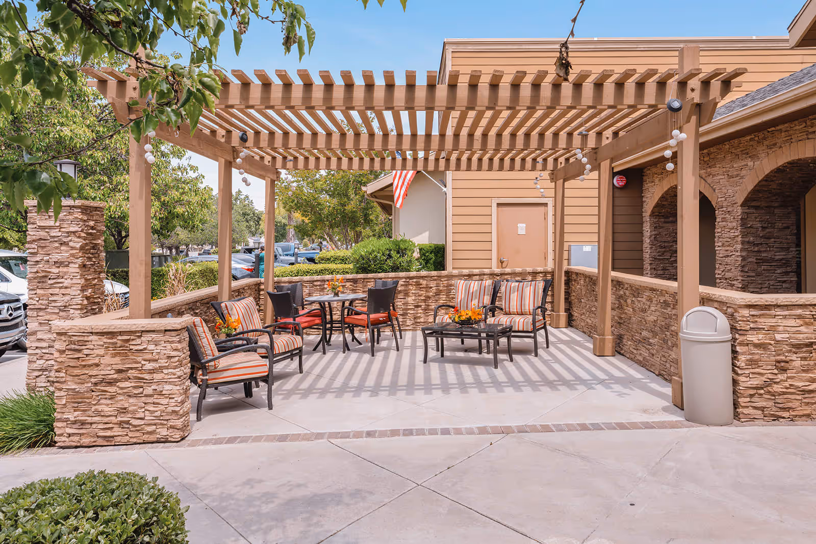 An outdoor patio area featuring a wooden pergola, comfortable seating with striped cushions, and small tables adorned with flowers. The backdrop includes lush greenery and a building facade.