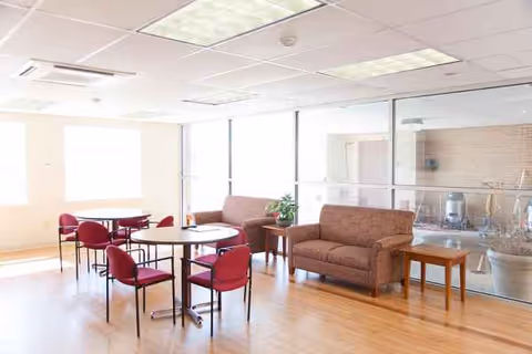 Bright indoor common area with round tables, red chairs, and beige sofas.