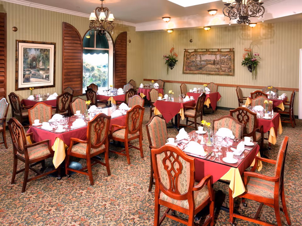Dining room with multiple tables set with red tablecloths, wooden chairs, chandeliers, and wall decorations.