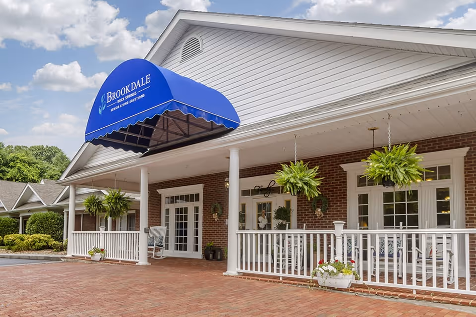 Front exterior view of Brookdale Rock Springs senior living facility with a blue awning displaying the facility name. The building has a brick facade, white columns, hanging plants, and a white railing enclosing a porch area with rocking chairs.
