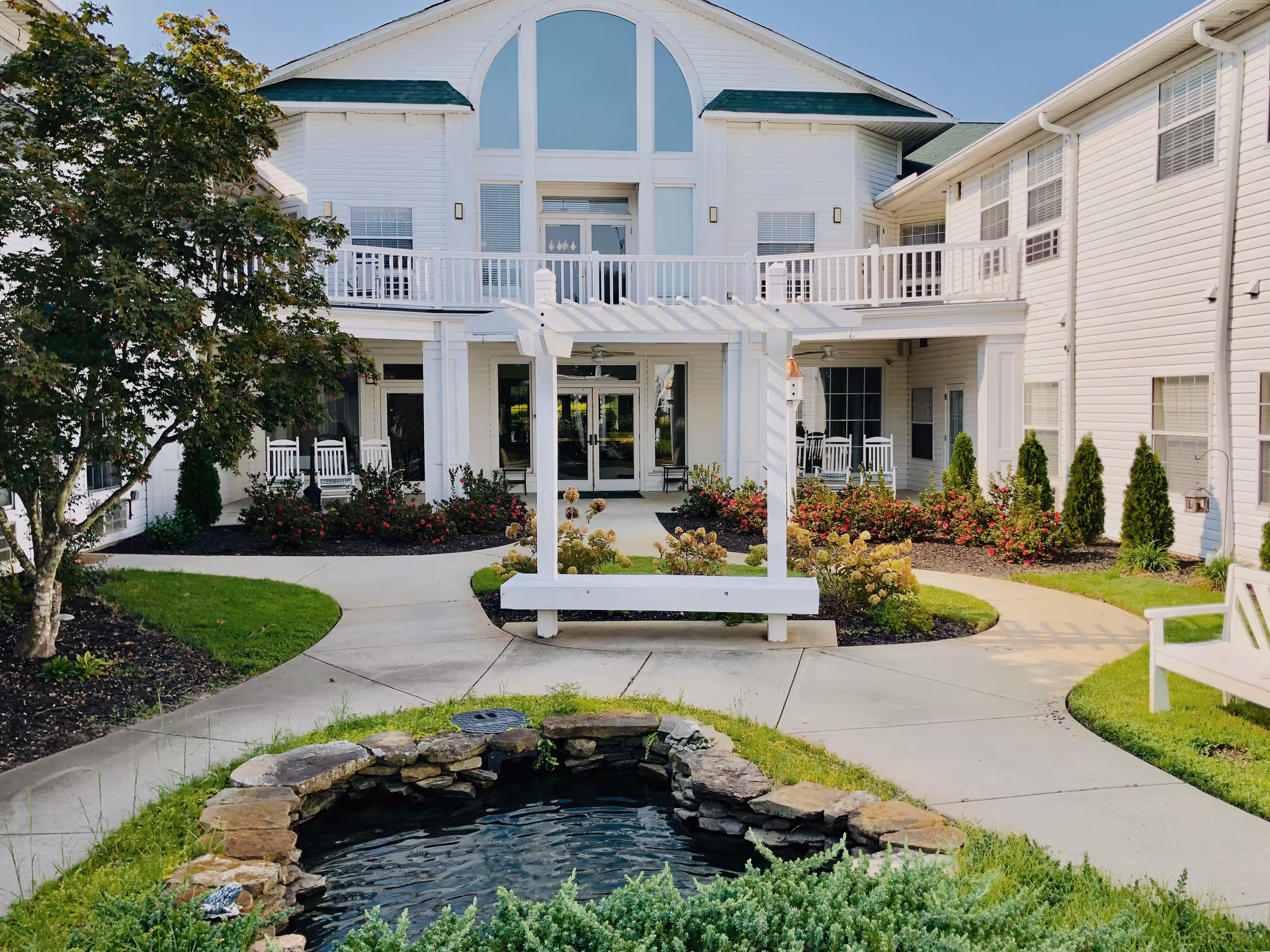 Front courtyard of a white senior living building with a small pond, pergola bench, walkways and landscaped gardens.