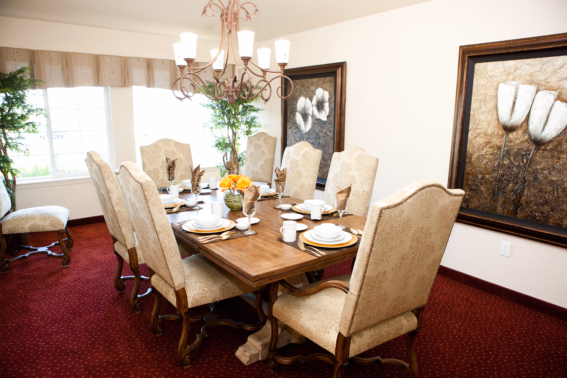 A formal dining room with a wooden dining table set for eight people. The table is adorned with plates, bowls, cups, gold-colored cutlery, and folded brown napkins in wine glasses. A floral centerpiece with orange flowers is in the middle of the table. The room has upholstered high-back chairs with a beige patterned fabric, red carpet flooring, two large framed floral paintings on the wall, and large windows with beige valances letting in natural light. A chandelier with multiple lights hangs above the table.