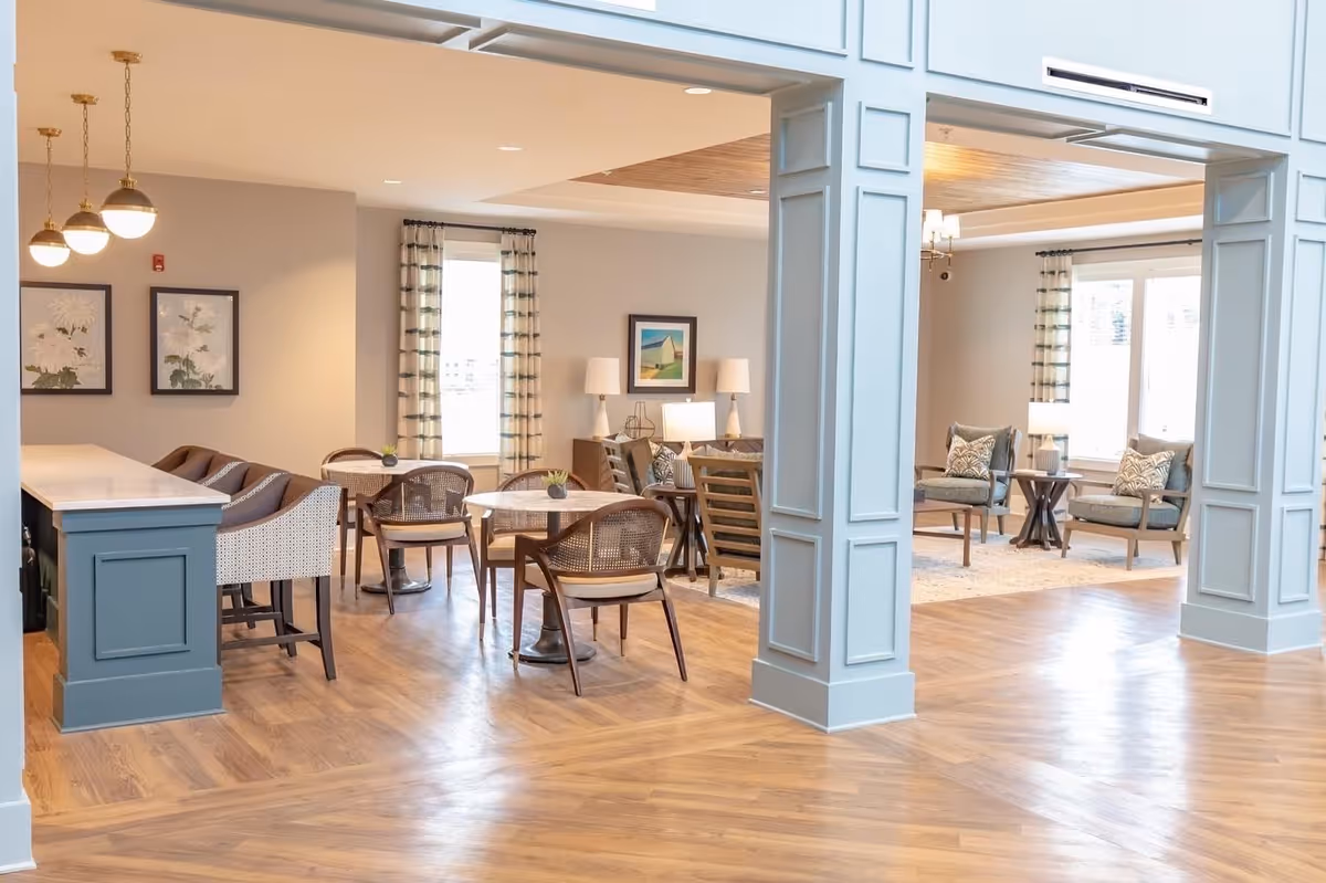 A bright and spacious common area in a senior living facility featuring wooden flooring, a seating area with armchairs and side tables, small round tables with chairs, a counter with bar stools, and decorative wall art and lamps.
