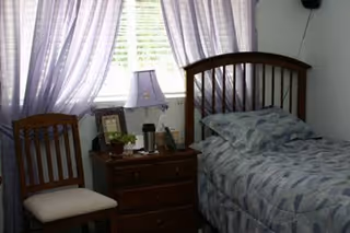 A bedroom with a wooden bed frame and a bed made with a blue patterned comforter and pillow. Next to the bed is a wooden nightstand with a lamp, framed photos, and other small items. A wooden chair with a cushioned seat is placed beside the nightstand. The window behind the nightstand has sheer white curtains allowing light to enter the room.
